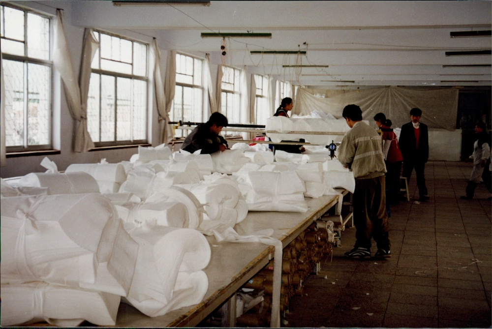 Manufacturing workers in China gathered around cutting tables with dozens of large stacks of white disposable fabric tied off in sections laying on the tables