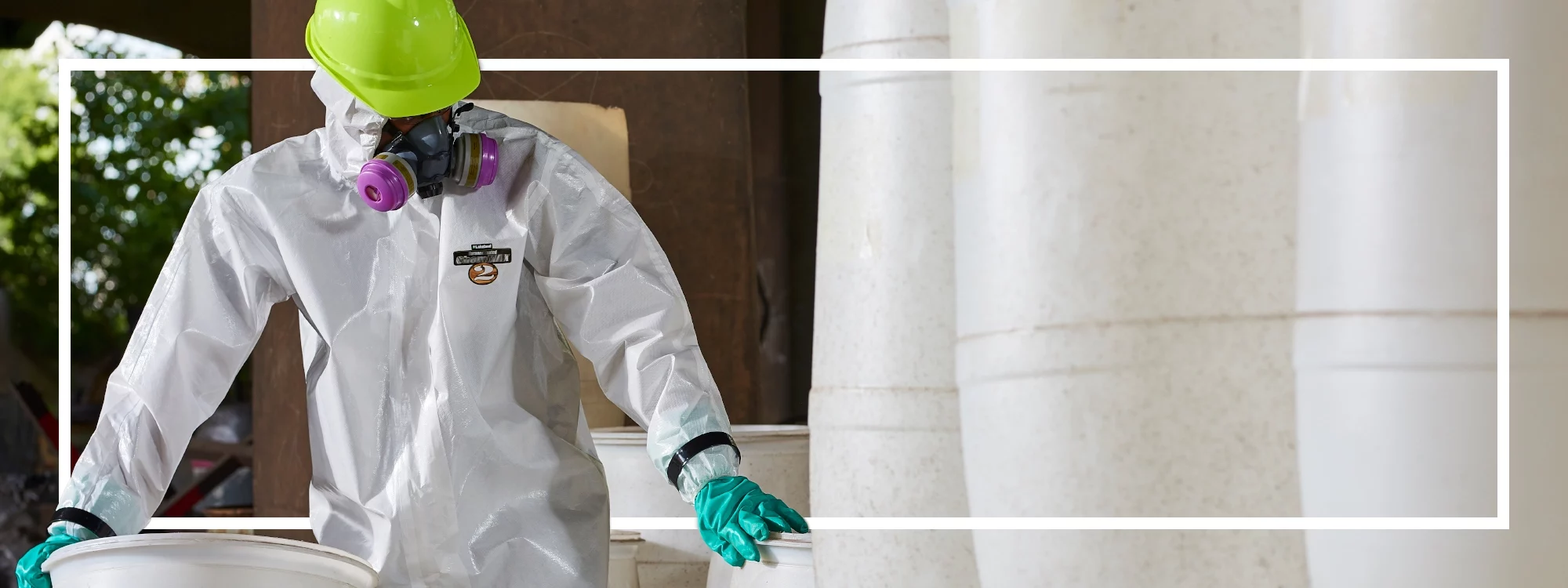 Un trabajador de una planta química que lleva Lakeland&#039;s ChemMax 2 y una mascarilla de respiración mueve bidones de plástico blanco de sustancias químicas desconocidas.