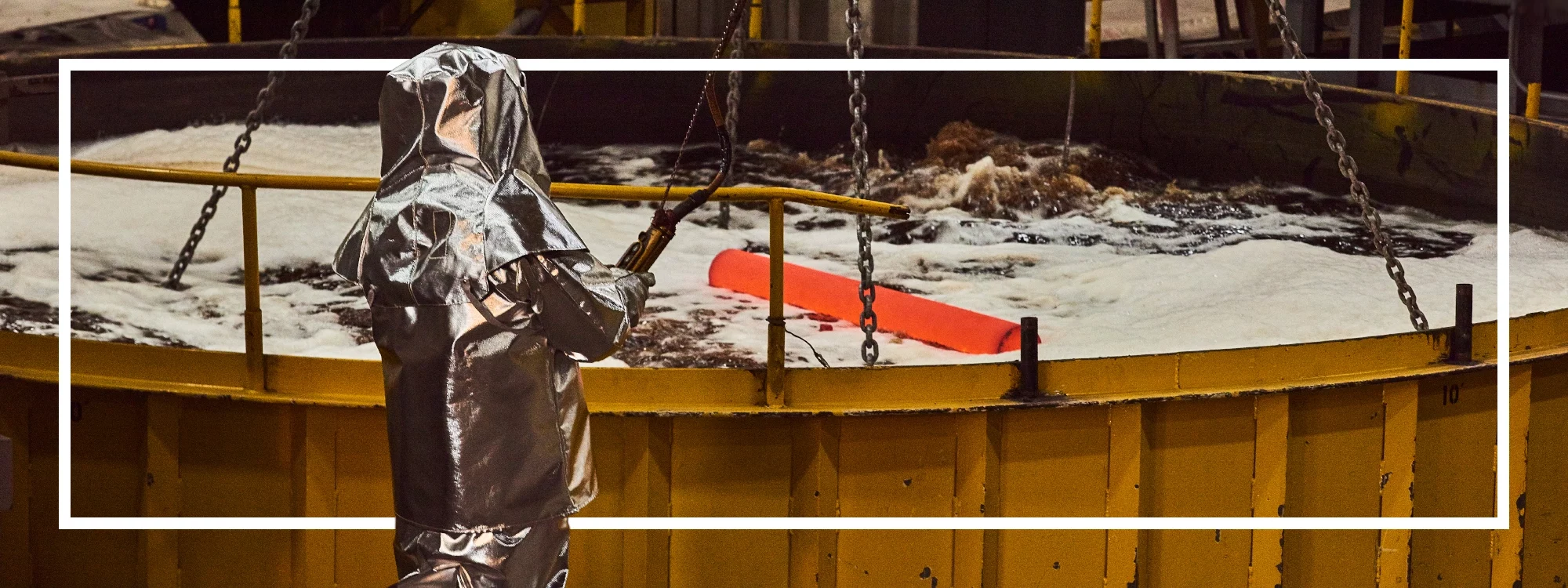 Worker wearing an aluminized garment lowers a cylindrical mass of hot, radiating metal into a large cooling tank