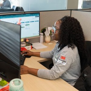 Woman working at dual monitors in office cubicle.